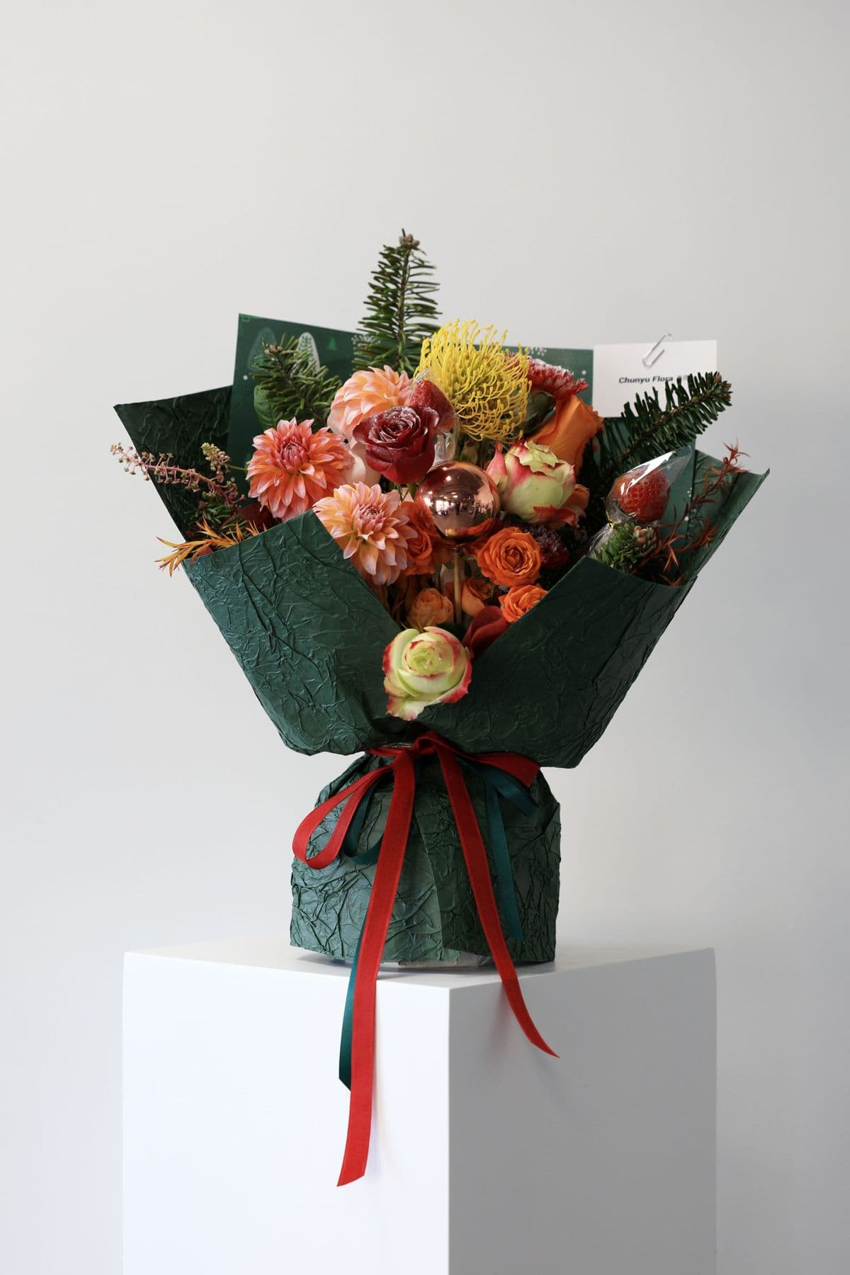 Hand-tied bouquet of orange roses and dahlias wrapped in dark green paper with a red ribbon